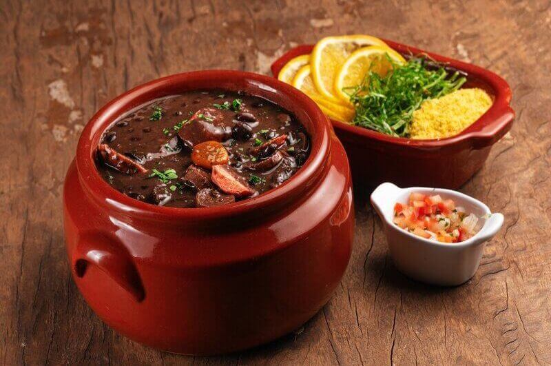 Recipiente de vidro marrom com feijoada e linguiça em cima de uma mesa de madeira com dois recipientes contendo laranja e tomate