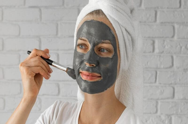 Mulher com toalha branca na cabeça, aplicando argila verde no rosto