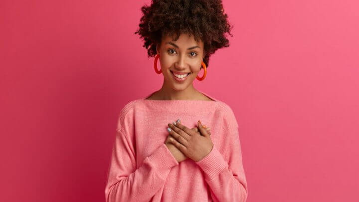 Mulher com blusa e brinco rosa com a mão no coração e sorrindo