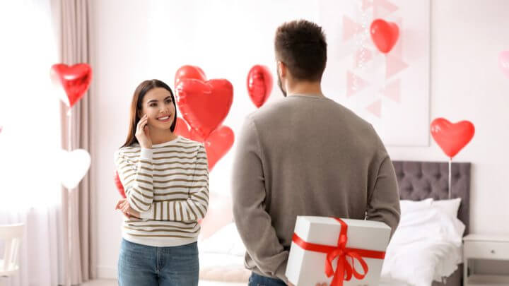 Homem segurando caixa surpresa nas costas para presentear mulher. O quarto está decorado de balões vermelhos.