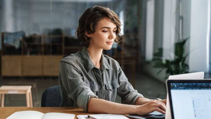 Mulher de cabelo curto e camisa cinza digitando em notebook