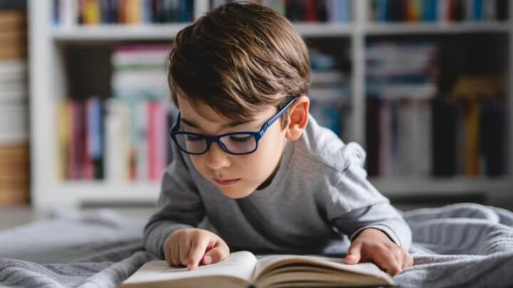 Menino usando óculos deitado lendo livro