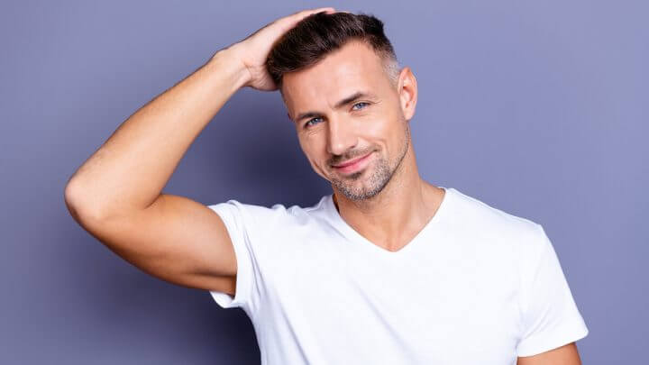 Homem sorrindo vestindo blusa branca e com as mãos no cabelo em frente a um fundo roxo