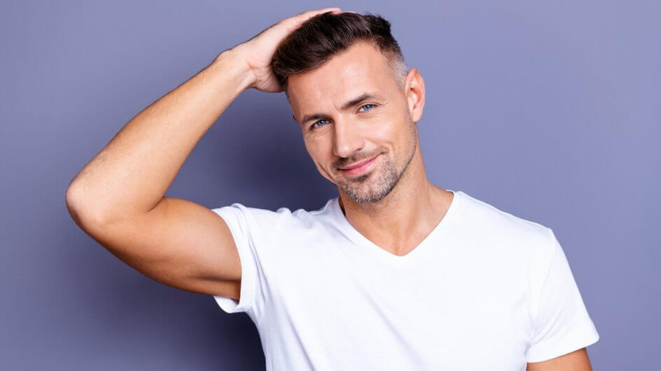 Homem sorrindo vestindo blusa branca e com as mãos no cabelo em frente a um fundo roxo