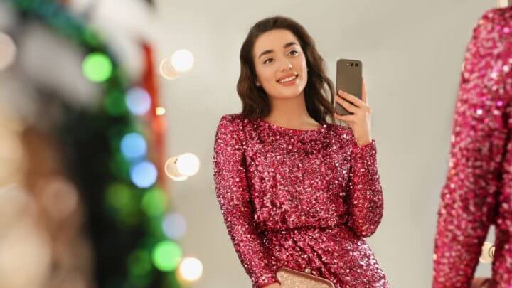 Jovem com vestido de lantejoulas rosa sorrindo e tirando foto no espelho