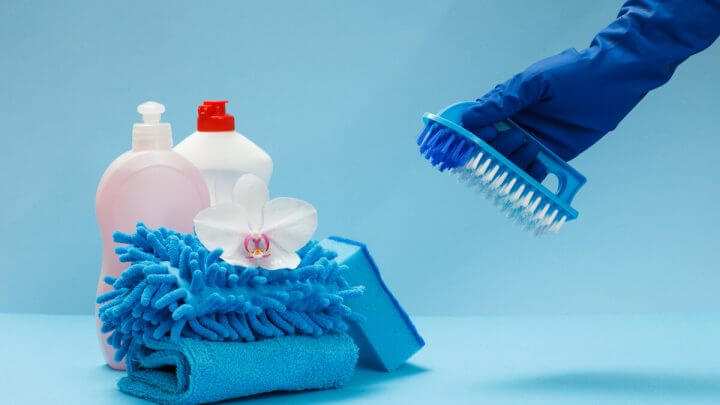 Produtos de limpeza em cima de um fundo azul com uma mão com luva azul segurando uma escova de lavar roupa