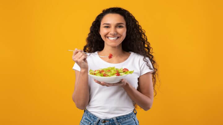 Mulher sorrindo e comendo salada em fundo amarelo.