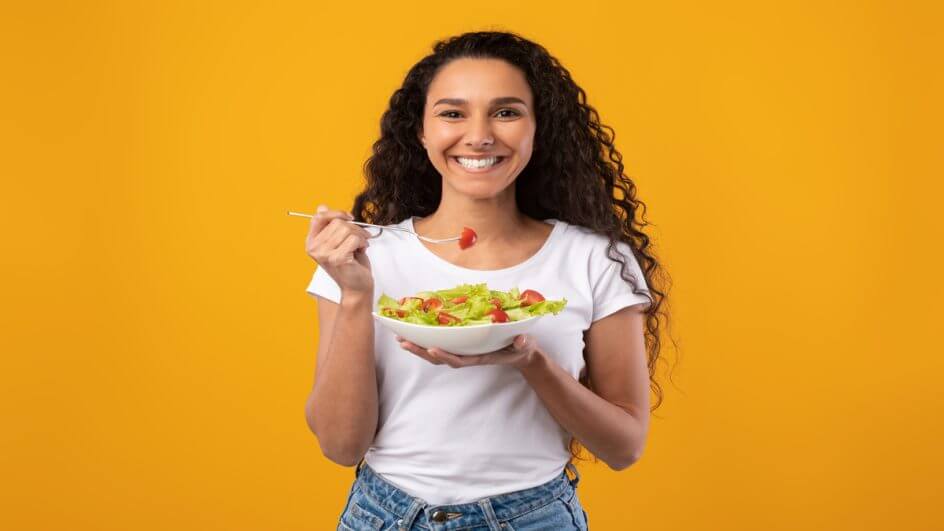 Mulher sorrindo e comendo salada em fundo amarelo.