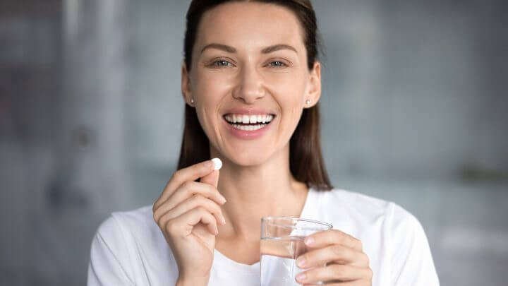 Mulher sorrindo segurando um copo com água e cápsula de remédio