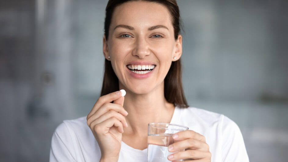 Mulher sorrindo segurando um copo com água e cápsula de remédio
